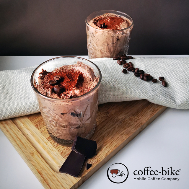 Two glasses of espresso chocolate mousse stand on a wooden board and are separated by a gray towel, on which beans are spread, right below the Coffee-Bike logo