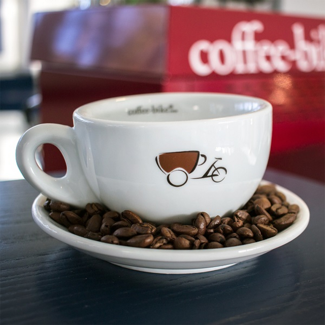 White cup with Coffee-Bike logo stands on saucer covered with coffee beans, in the background blurred portafilter machine