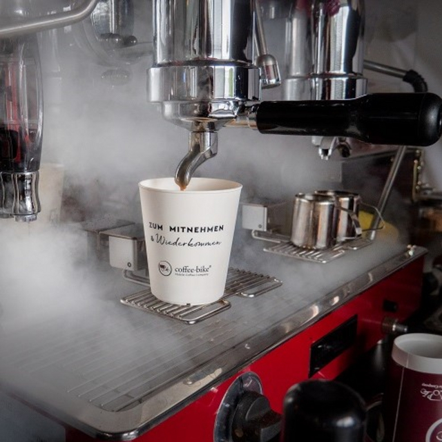 Weißer Pfandbecher mit der schwarzen Aufschrift "Zum Mitnehmen und Wiederkommen" und dem Coffeee-Bike Logo steht unter der Siebträgermaschine im Dampf