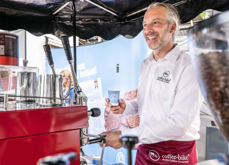 Barista mit Coffee-Bike Dienstkleidung hält hinter der roten Kaffeemaschine einen Kaffeebecher mit Firmenlogo in der Hand