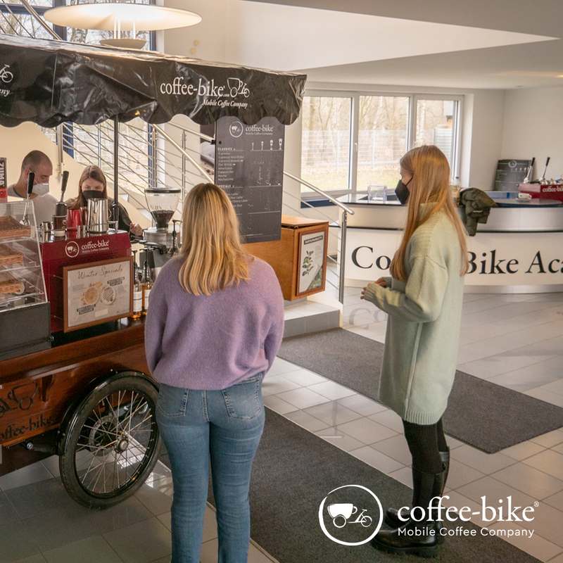 A woman with purple sweater and a woman with gray sweater are standing facing the Coffee-Bike, behind which two people are standing, white Coffee-Bike logo on the lower left side
