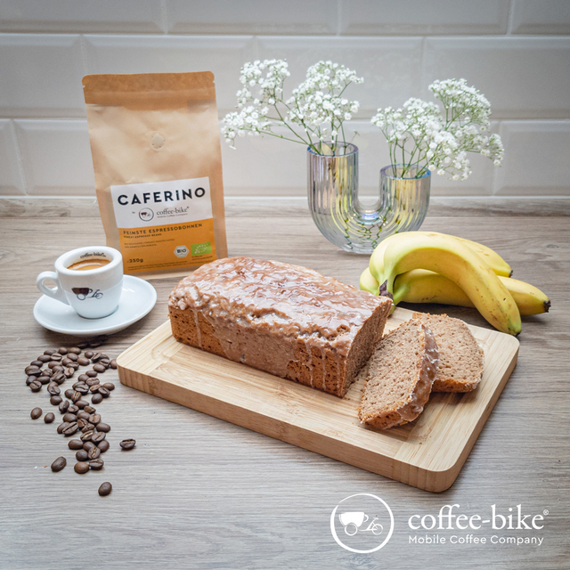 Coffee-Bike espresso cup stands in front of a Caferino bean packet, next to it flowers, bananas and a cut espresso banana bread