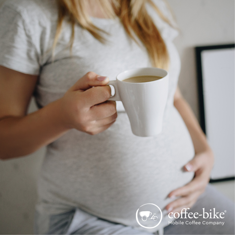 Schwangere Frau mit Hand auf dem Bauch und einer Kaffeetasse in der anderen Hand, thematisiert Kaffee in der Schwangerschaft
