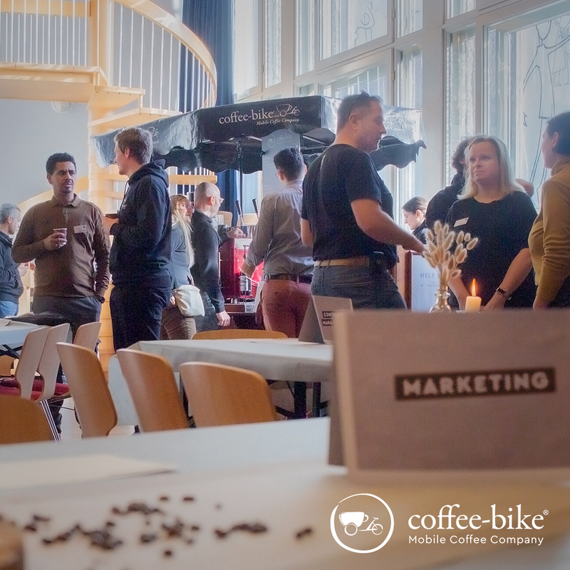 [Translate to UK:] In the background, people stand in front of and next to a Coffee-Bike; in the foreground, a table is decorated with beans and a sign.