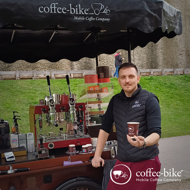 A man in Coffee-Bike clothing stands in front of the bike with a cup in his hand
