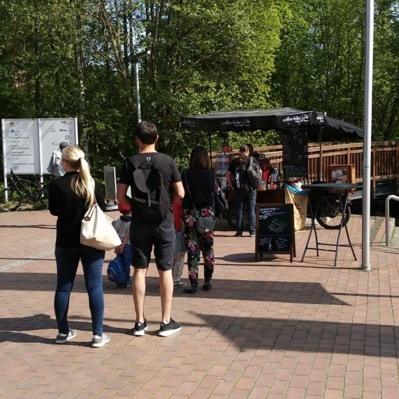 Several people line up at the Coffee-Bike, which is in front of a wooden bridge and trees.