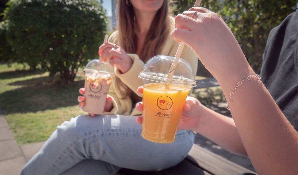 In the foreground a Coffee-Bike Clearcup is held with orange juice, in the background sits a woman with denim pants and light jacket stirring an Iced Latte in the Clear Cup with a straw, bottom right white Coffee-Bike logo