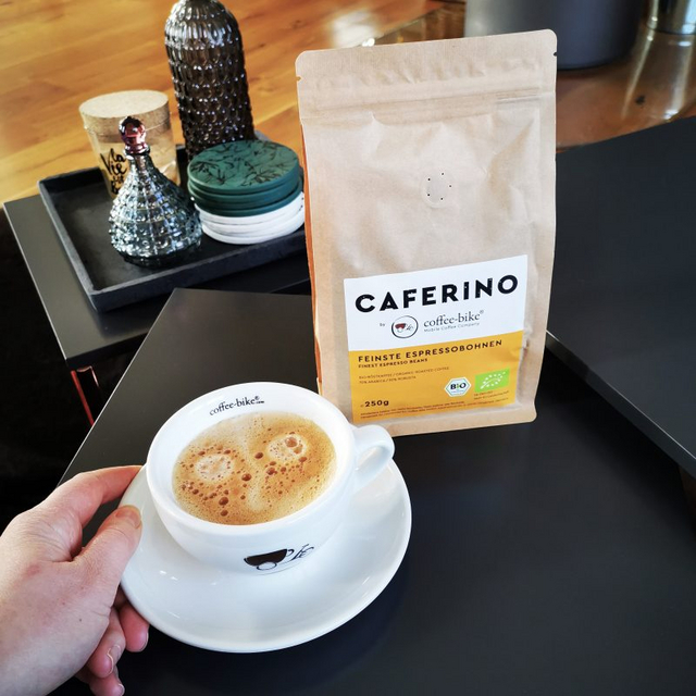 Eine Cappuccinotasse mit Coffee-Bike Logo und eine Caferino Espressopackung stehen auf einem schwarzen Tisch, während an der Tasse eine linke Hand liegt und im Hintergrund vier Dekoartikel stehen