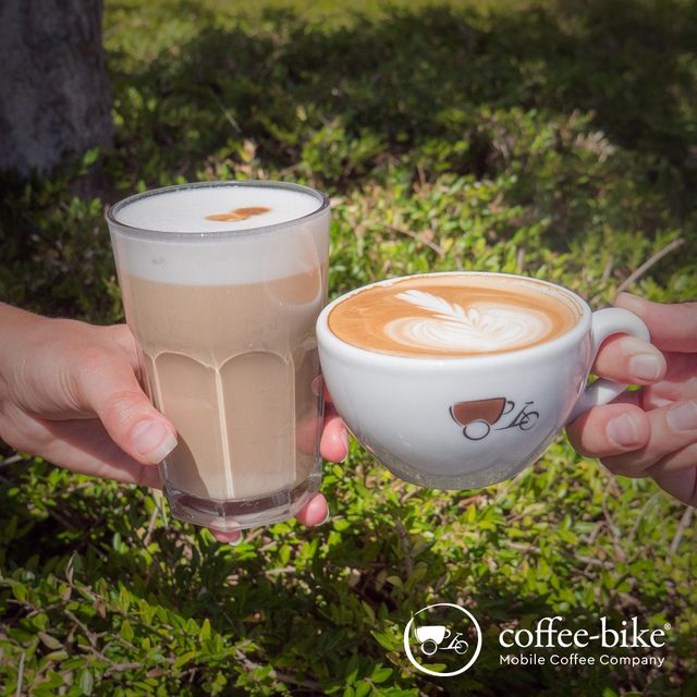 Zwei Hände stoßen von beiden Seiten mit einem Latte Macchiato Glas und einer weißen Coffee-Bike Cappuccinotasse vor einem naturgrünen Hintergrund an, unten rechts weißes Coffee-Bike Logo