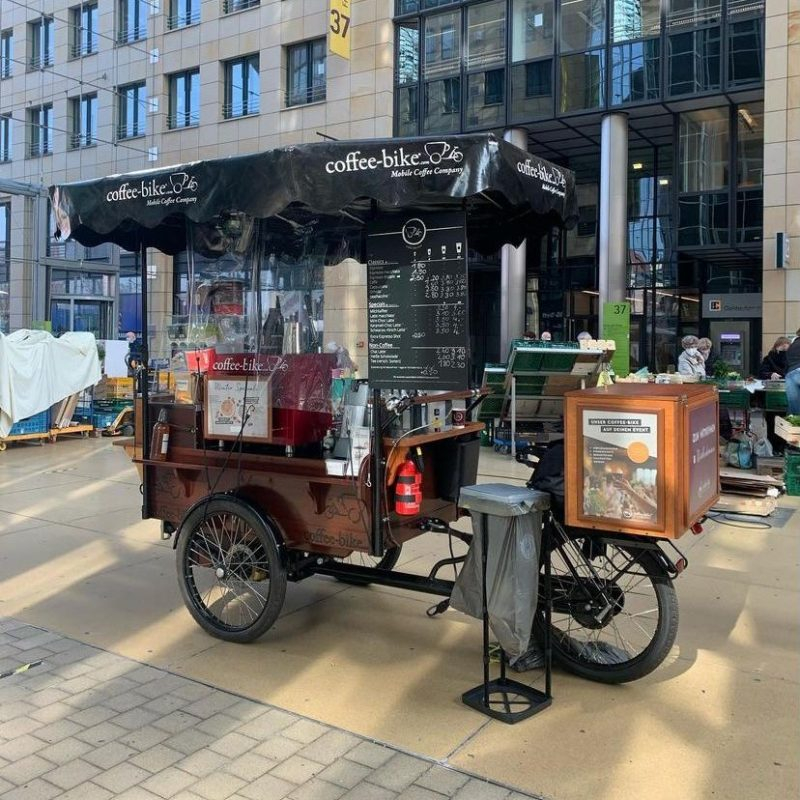 A fully equipped Coffee-Bike stands in front of a building with a bright frontage