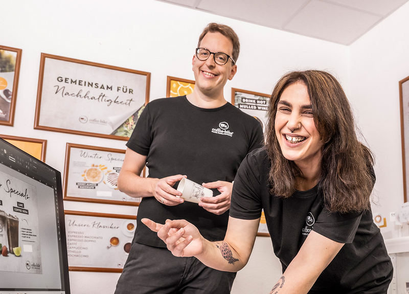 Zwei Osnabrücker Mitarbeiter zeigen in einem Büroraum auf den Bildschirm und lachen
