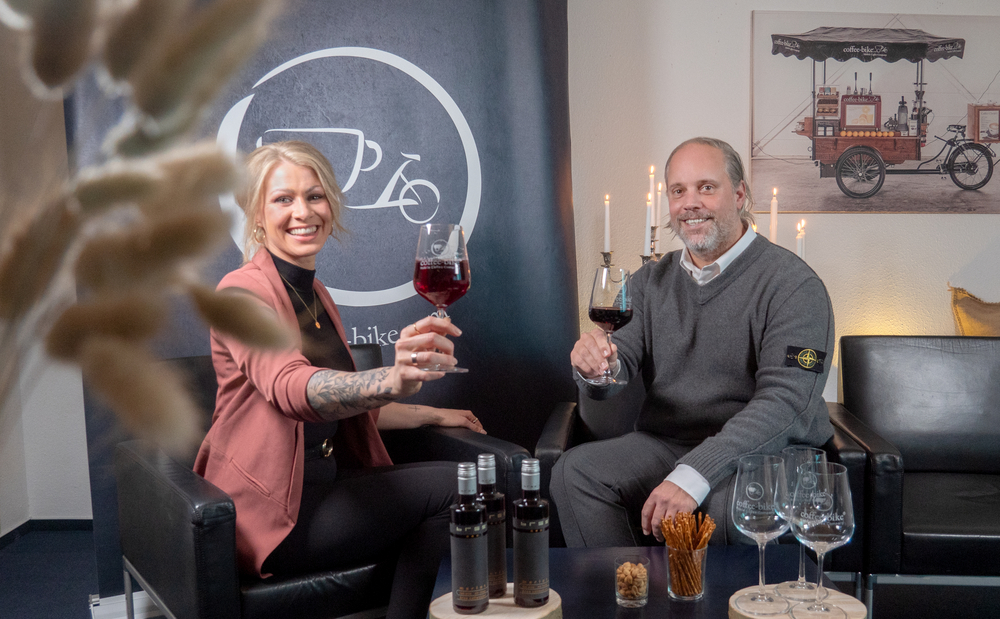 A woman with pink blaser and a man with gray sweater hold up a red wine glass, below red wine bottles and glasses, Coffee-Bike logo behind the woman and below right