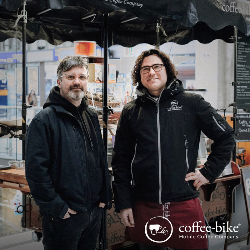 Zwei Männer stehen vor einem Coffee-Bike, unten rechts weißes Coffee-Bike Logo