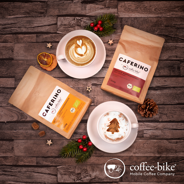 Auf braunem Hintergrund liegen eine Caferino Espressobohnenpackung, eine Kaffeebohnenpackung, zwei Tassen mit Untertassen mit Herz Latte Art und Tannenbaum Latte Art, Weihnachtsdeko und weißes Coffee-Bike Logo unten rechts