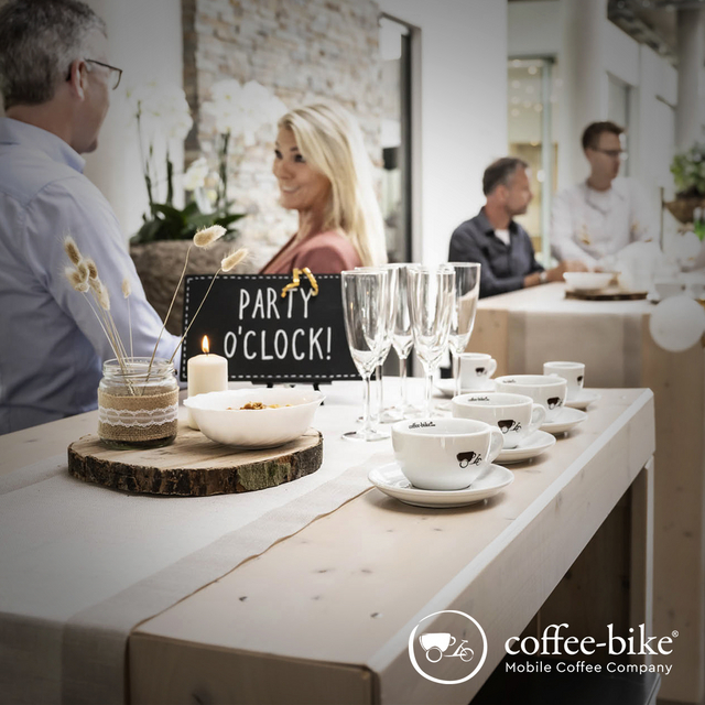 Auf Holztischen stehen Coffee-Bike Tassen, Sektgläser, Dekoration und ein schwarzes Schild mit den Worten Party O Clock, dahinter stehen sich unterhaltende Menschen, unten rechts weißes Coffee-Bike Logo
