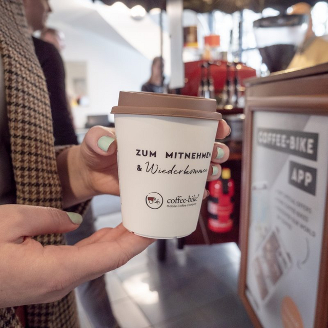 Ein Mehrwegbecher auf dem in schwarzer Schrift "Zum Mitnehmen und Wiederkommen" steht, wird von zwei Händen gehalten, im Hintergrund ist ein unscharfes Coffee-Bike