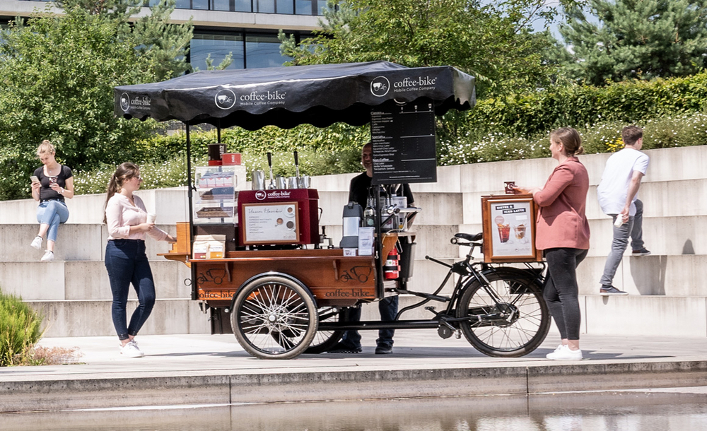 Vollausgestattetes Coffee-Bike steht vor Betontreppen und grünen Büschen, daneben Personen, die Kaffee trinken