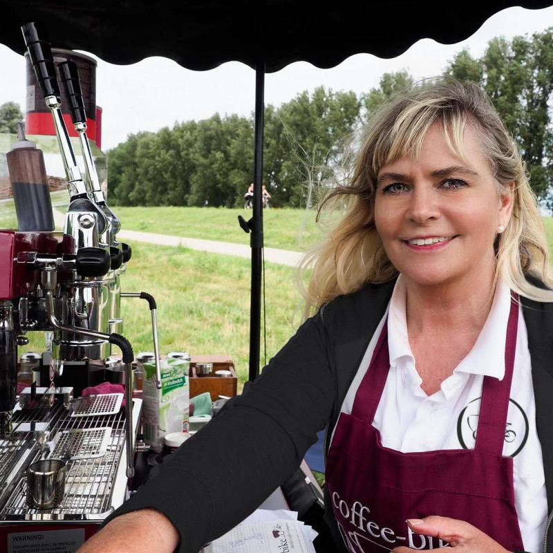 Eine Frau mit weißer Bluse, roter Schürze und schwarzer Jacke steht neben der Siebträgermaschine eines Coffee-Bikes auf einer grünen Wiese.