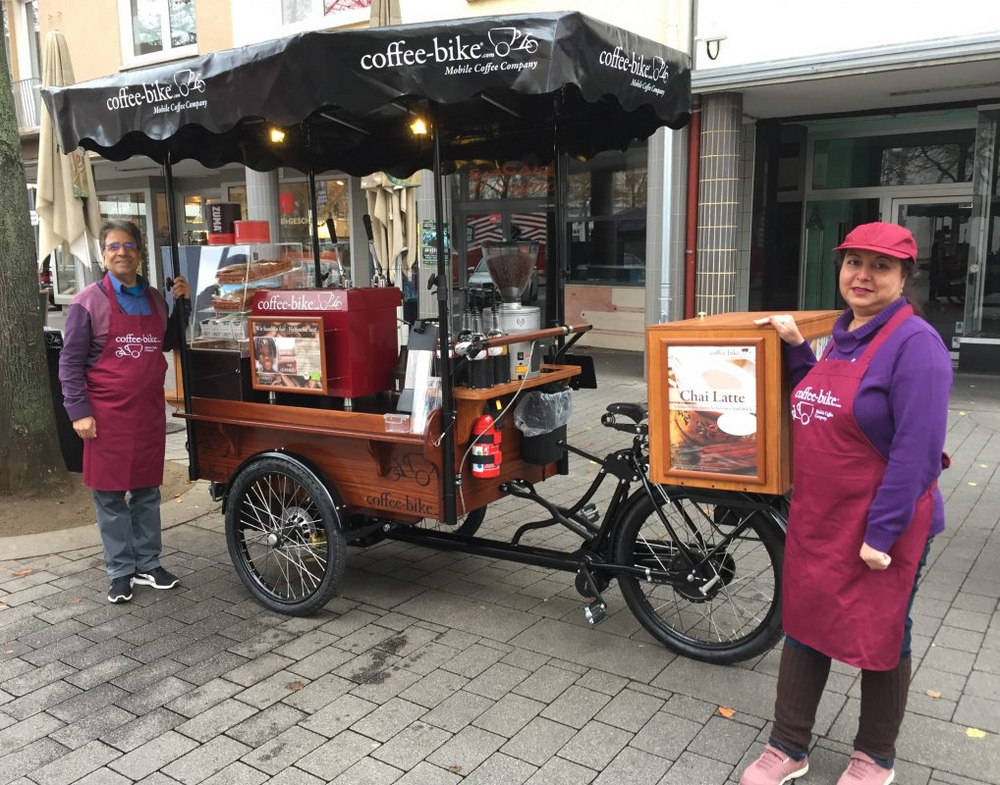 In a downtown area, a man wearing a Coffee-Bike apron is standing to the left of the Coffee-Bike and a woman wearing an apron is standing to the right of it, he is holding a side bar with his left hand, her hand is on the transport box