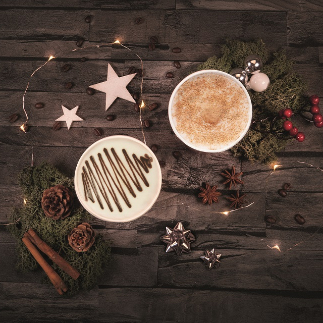 A specialty coffee with chocolate sauce and one with cinnamon topping stand on a dark background adorned with a string of lights and Christmas decorations.