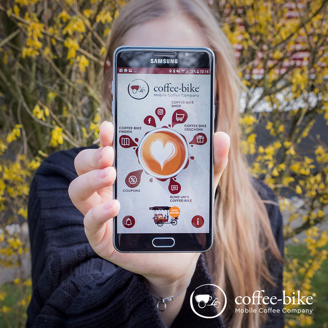 Eine Frau mit blonden Haaren hält ein Samsung Smartphone mit der rechten Hand vor ihr Gesicht, auf dem Display ist die Coffee-Bike App geöffnet, unten rechts weißes Coffee-Bike Logo