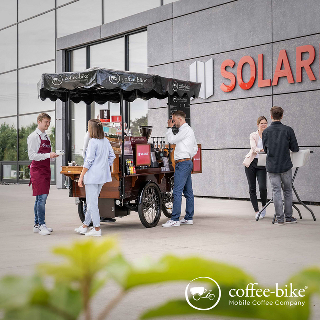 Customers are standing in front of a fully equipped Coffee-Bike, on the left is the barista wearing a white shirt and red Coffee-Bike apron