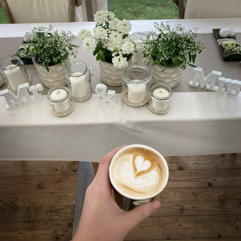 Eine linke Hand hält einen Cappuccino mit Latte Art Herz vor einen für eine Hochzeit dekorierten Tisch mit Kerzen, Blumen und Mr und Mrs Aufstellern.