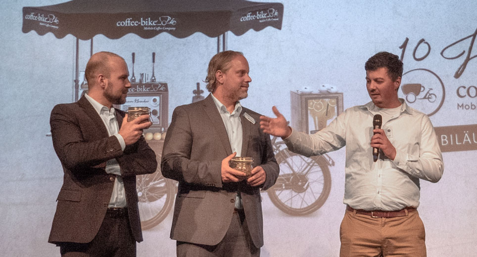 Mark Rüter, managerul Coffee-Bike, și fondatorul Tobias Zimmer vorbesc cu un partener de franciză pe o scenă în timpul unei prezentări a companiei.