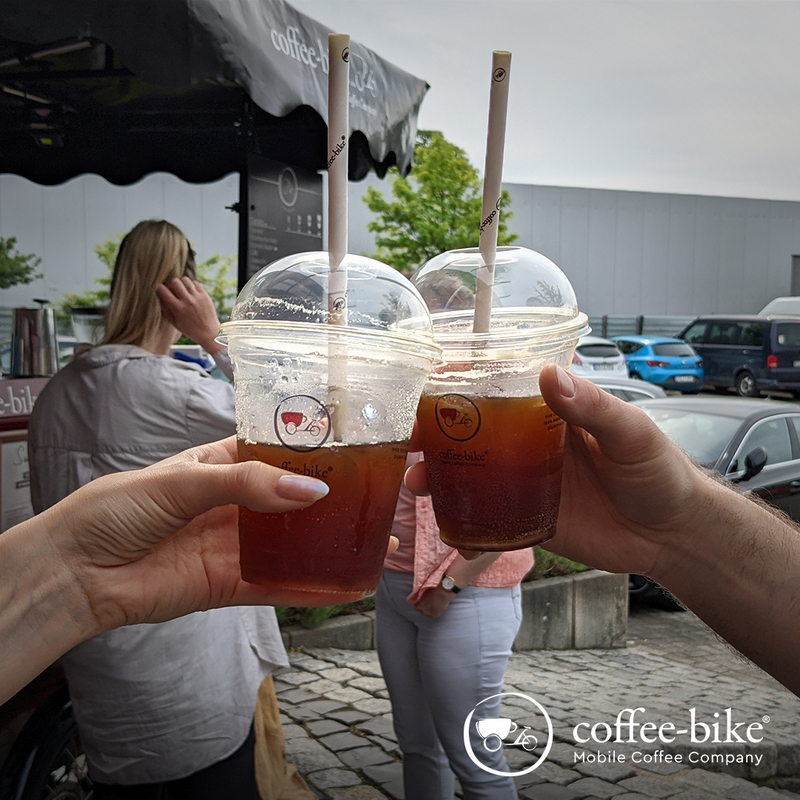 Two hands push from both sides with the Coffee-Bike clear cups, in which is up to half a brown liquid, in the background are cars and a woman and cut the Coffee-Bike, bottom right white Coffee-Bike logo