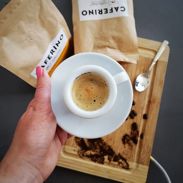 An espresso cup is held over a wooden board on which are beans, a spoon and two Caferino packets.