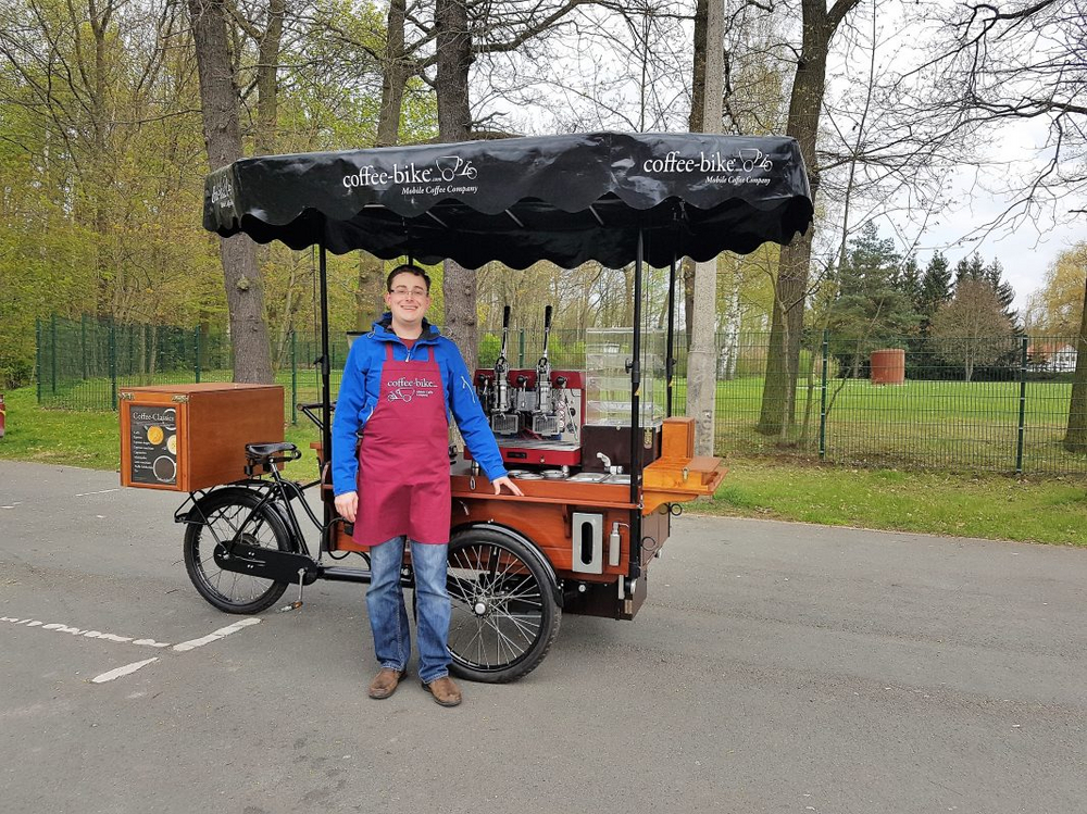Outside, a man in a blue jacket and red Coffee-Bike apron stands in front of a fully equipped Coffee-Bike