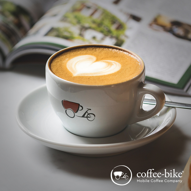 Eine weiße Tasse mit Coffee-Bike Logo beinhaltet einen Cappuccino mit Latte Art, dahinter ist eine aufgeschlagene Zeitschrift, rechts unten das weiße Coffee-Bike Logo