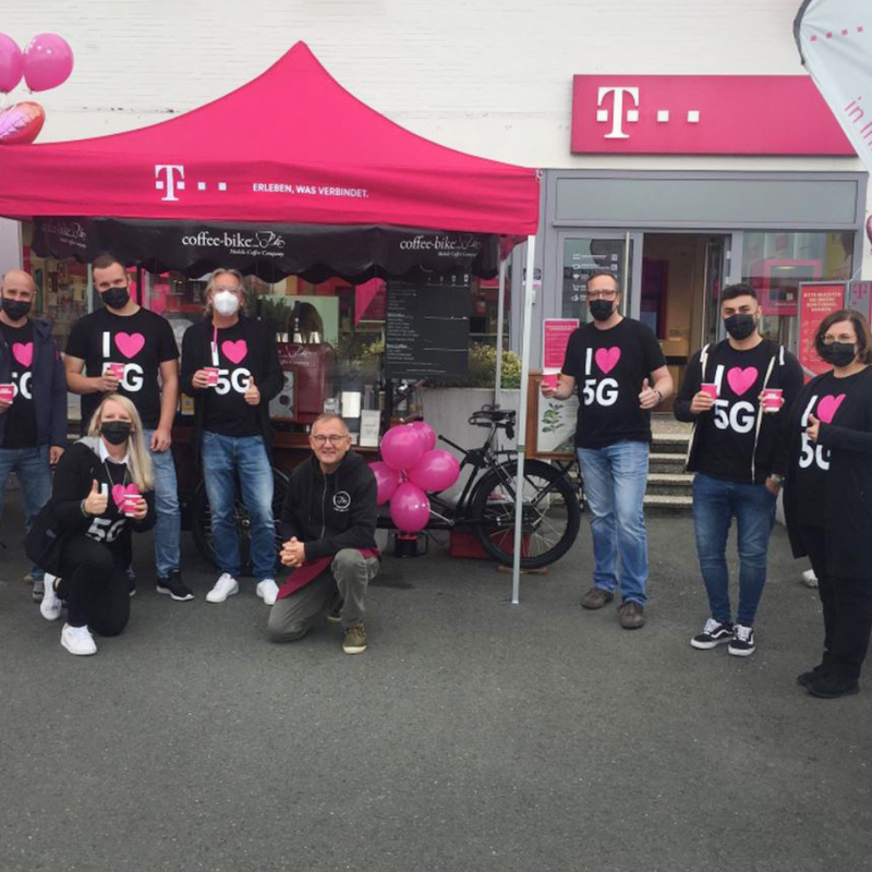 The Coffee-Bike is in the background in front of a Telekom store under a pink Telekom pavilion, employees are standing in front of and next to it, the Coffee-Biker is squatting in the middle