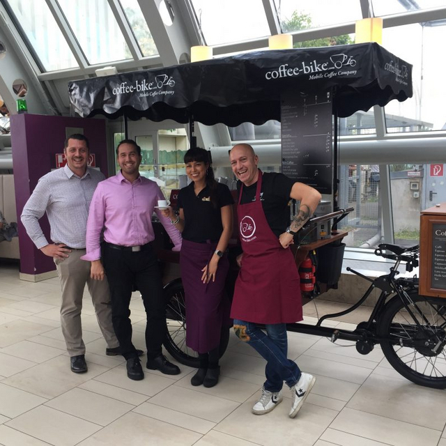 A Coffee-Biker stands with three other people in front of the Coffee-Bike in the KaDeWe.