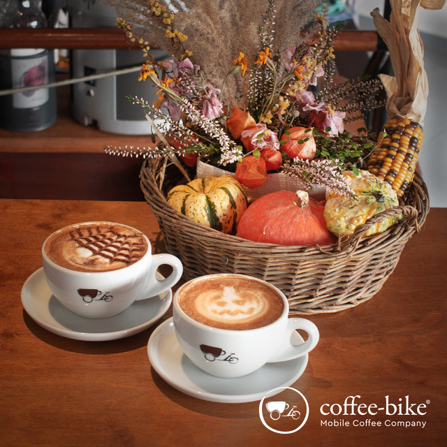 Two white coffee bike cups with spider web and pumpkin latte art stand in front of a basket with different kinds of pumpkins and autumn decorations, bottom right white Coffee-Bike logo