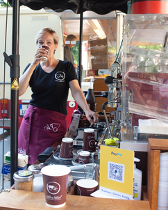 A barista with black Coffee-Bike shirt and red apron drinks a coffee behind a Coffee-Bike, in the foreground is a Coffee-Bike mug
