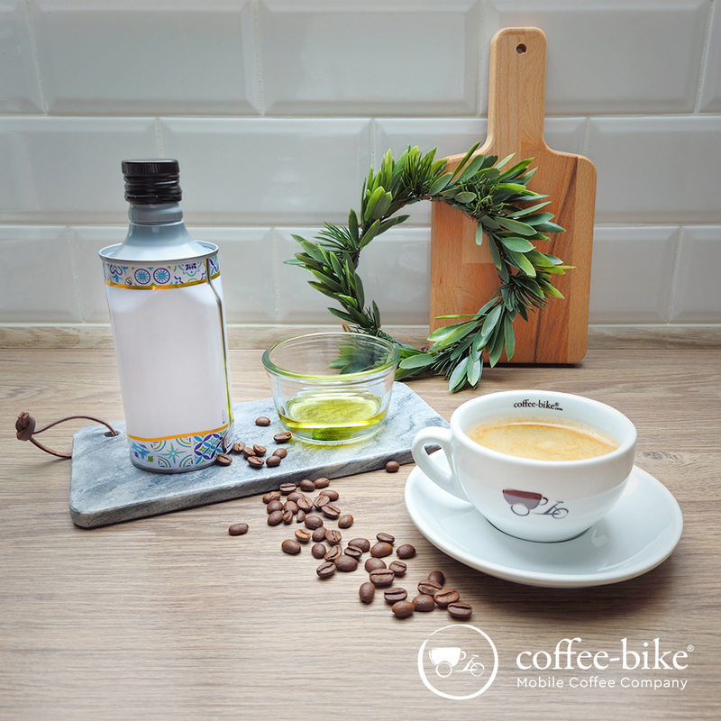 Gebrandete Kaffeetasse steht vor einem Brettchen, einem Olivenblattkranz, einer Flasche und einem Glas mit Olivenöl. Daneben liegen Kaffeebohnen.