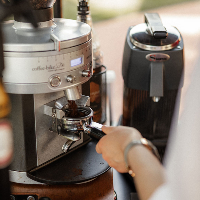 Eine linke Hand hält den Siebträger unter die Mühle, aus der gemahlenes Kaffeepulver kommt
