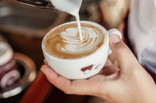 Barista gießt Latte Art Herz mithilfe eines Milchkännchens in eine weiße Cappucinotasse aus Porzellan mit Coffee-Bike Logo