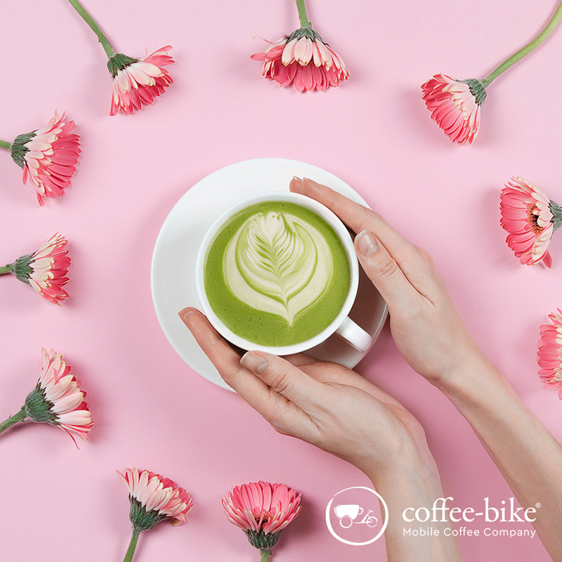 On pink background is a white cup with green coffee surrounded by two hands and several pink flowers.