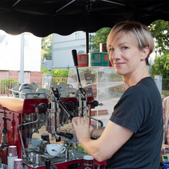 A barista pushes down the lever of the portafilter machine, underneath is a white Coffee-Bike cup, she looks over her left shoulder