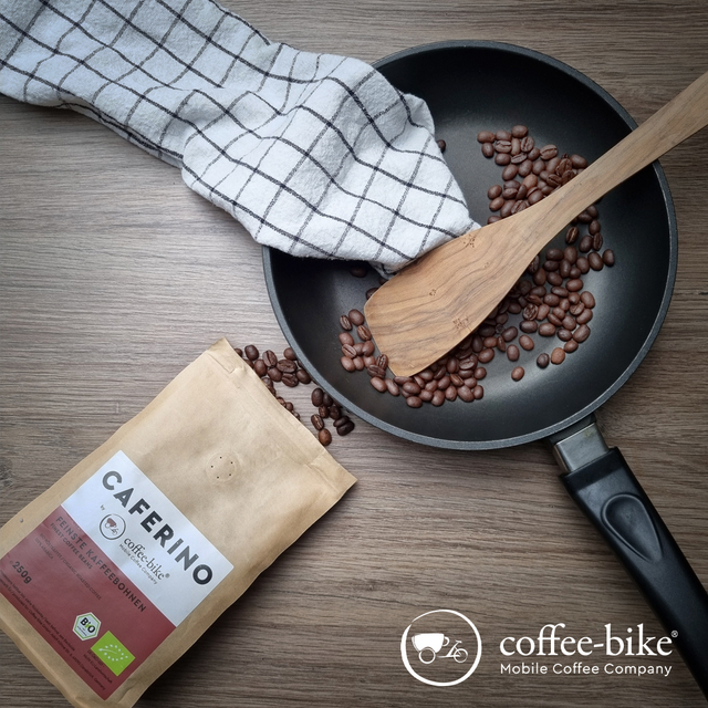 In a black pan are coffee beans, a wooden cooking spoon and a black and white towel, in front of it is an open Caferino coffee beans package