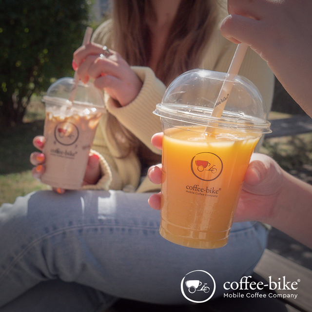 In the foreground a Coffe-Bike Clearcup with orange juice is held, in the background sits a woman with jeans trousers and bright jacket, who stirs an Iced Latte in the Clearcup with the straw, below right white Coffe-Bike logo