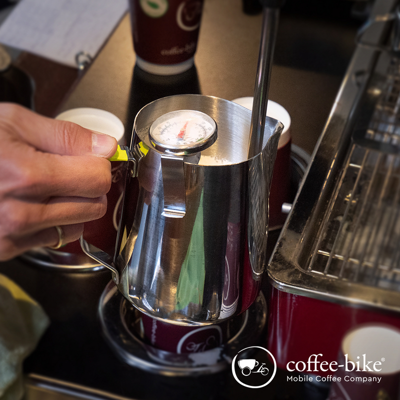 Eine rechte Hand hält eine Edelstahlkanne mit Milch und einem Thermometer unter die Dampflanze, rechts unten weißes Coffee-Bike Logo