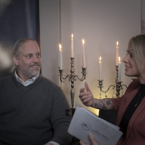 Im Hintergrund stehen zwei Kerzenleuchter mit weißen brennenden Kerzen, davor sitzen links ein lächelnder Mann und rechts eine Frau mit Karten in der linken Hand