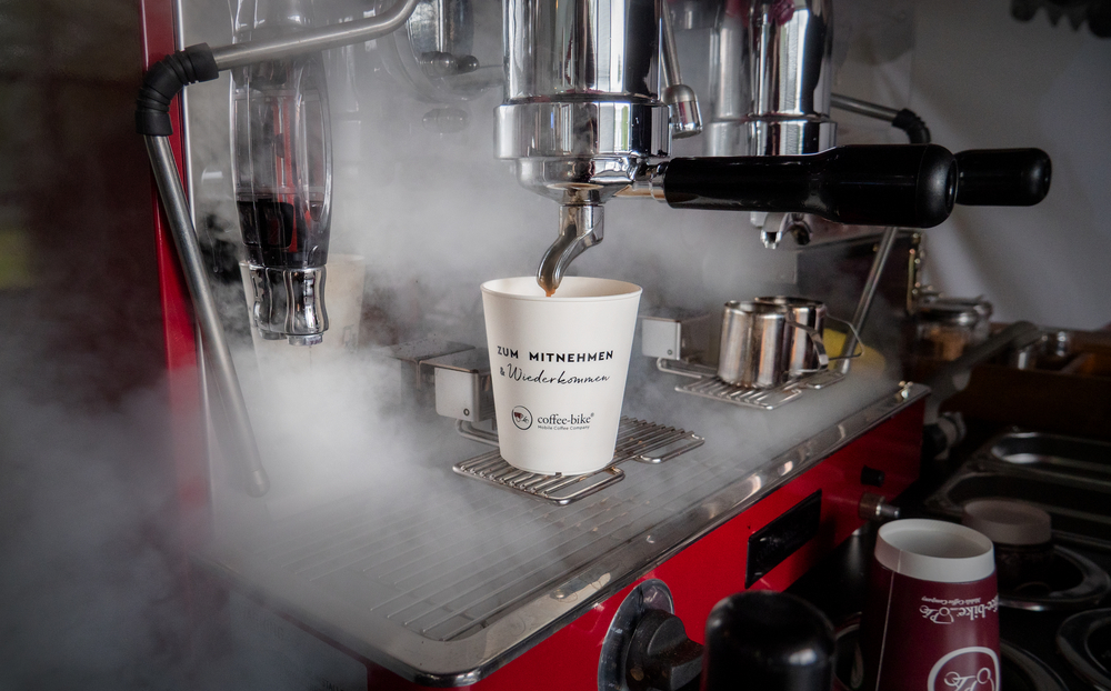 White deposit cup with the black inscription "Zum Mitnehmen und Wiederkommen" and the Coffee-Bike logo is under the portafilter machine in the steam