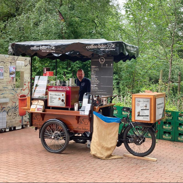 Barista steht auf einem gepflasterten Platz hinter einem voll ausgestatteten Coffee-Bike, das umgeben von Bäumen ist.