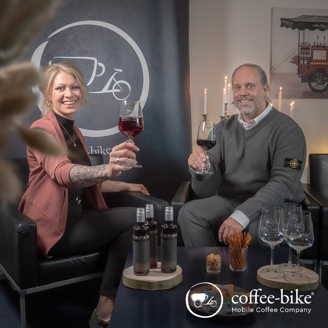 Eine Frau mit rosa Blaser und ein Mann mit grauem Pullover halten ein Rotweinglas hoch, darunter Rotweinflaschen und Gläser, Coffee-Bike Logo hinter der Frau und unten rechts