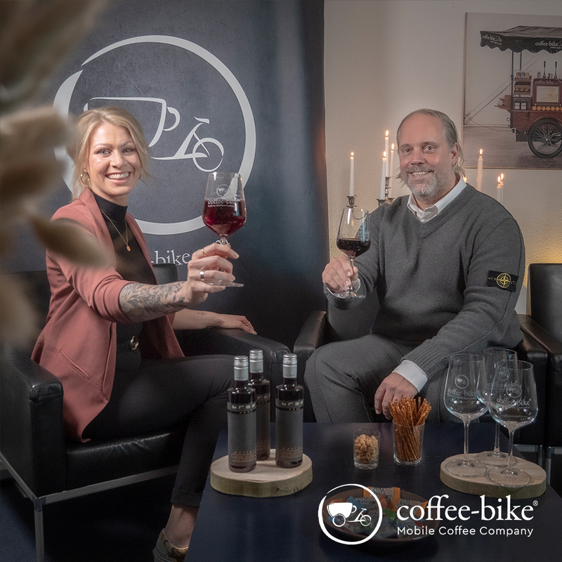 A woman with pink blaser and a man with gray sweater hold up a red wine glass, below red wine bottles and glasses, Coffee-Bike logo behind the woman and below right