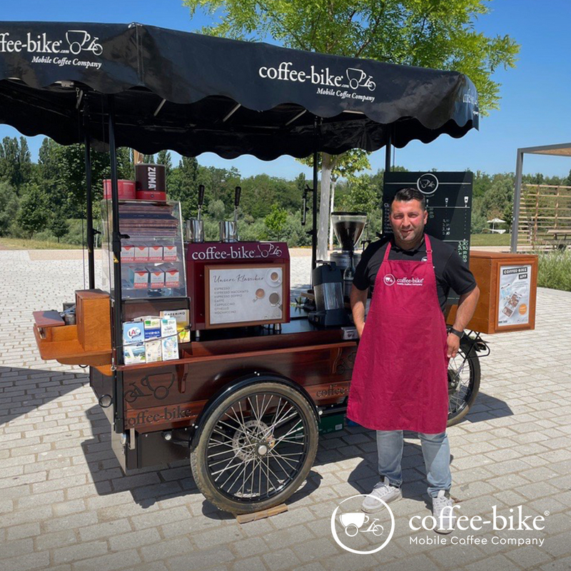 Ein Barista mit schwarzem Hemd und roter Schürze steht rechts vor einem vollausgestatteten Coffee-Bike bei blauem Himmel, unten rechts weißes Coffee-Bike Logo